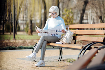 Important news. Serious aged woman reading a newspaper while sitting on the bench