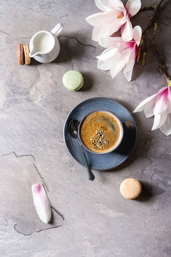 Blue cup of black espresso coffee with french dessert macaroons, cream and spring flowers magnolia branches over grey texture background. Top view, space. Spring greeting card