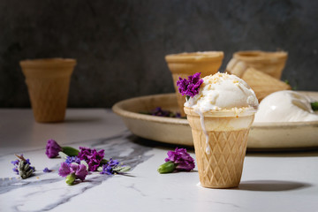 Obraz na płótnie Canvas Homemade vanilla ice cream in small waffle cup served with purple edible flowers and metal spoon in ceramic plate on white marble kitchen table.