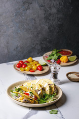 Variety of vegetarian corn tacos with vegetables, green salad, chili pepper served on ceramic plate with tomato and cream sauces with ingredients above on white marble kitchen table