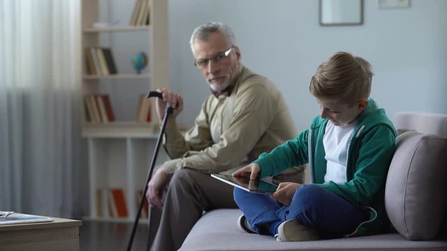 Boy playing video game on tablet, paying no attention at lonely grandfather