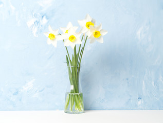 Fototapeta na wymiar Bouquet of fresh flowers, daffodils in vase in middle of table, opposite blue textured wall. Empty space for text.