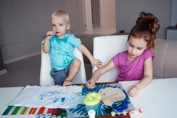 Two happy children blond boy and girl drawing paints and brushes and sitting at table at home.