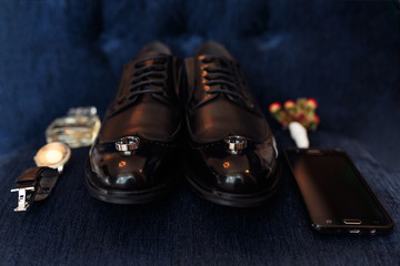 Accessories of a groom: shoes, boutonniere, phone, parfume and clock on dark blue textile surface. Wedding preparation