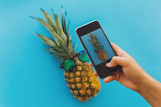 hand holding phone and taking photo of pineapple in stylish sunglasses on blue trendy paper background, flat lay. instagram blogging workshop concept. modern summer image