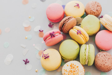 delicious colorful macaroons on trendy pastel gray paper with lilac flowers and confetti, flat lay. tasty pink, yellow, green and brown macaroons. space for text. candy for party