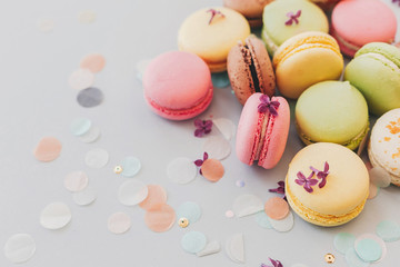 tasty pink, yellow, green and brown macaroons on trendy pastel gray paper with lilac flowers and confetti. space for text. delicious colorful macaroons candy for party. yummy background
