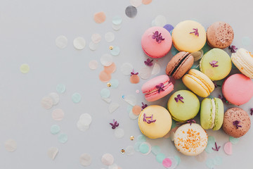 delicious colorful macaroons on trendy pastel gray paper with lilac flowers and confetti, flat lay. tasty pink, yellow, green and brown macaroons. space for text. candy for party