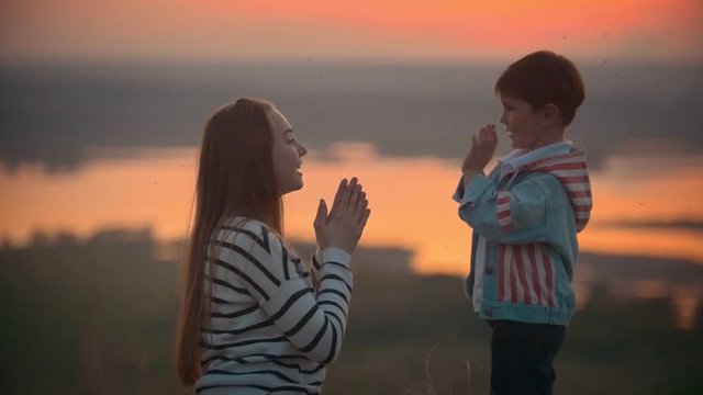 Young woman with little brother at summer meadow playing clap hands at sunset