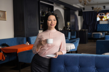 Nice business woman standing at table at restaurant and having coffee. Looking aside.