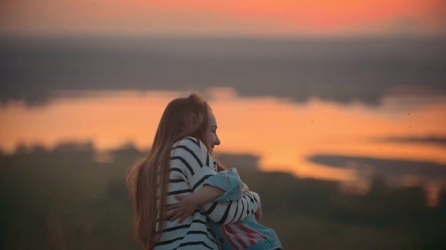 Warm summer holiday, young mom sitting in the meadow watching the sunset, in the background the river, her little son resorts to her and hugs, they laugh