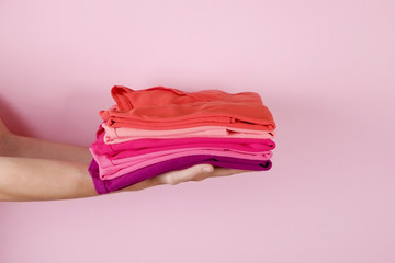 Close up of young woman hands holding stack of perfectly folded multicolor shirts. Female w/ pile of different color clothing in her arms on pale pink background. Laundry concept.