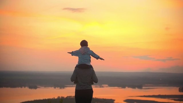 Mother and her little son have fun against the scarlet sunset .Mother holds child on shoulders,the child waves his hands and depicts a bird