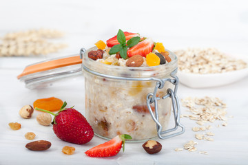 Oatmeal porridge in glass jar with fresh strawberries, nuts and dried fruit. Decorated with mint leaves, white table. 