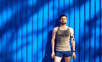 A man with a beard athlete runner in headphones against a blue wall background with space for text.