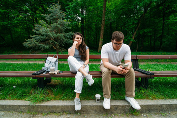 couple sitting on bench in city park. woman is boring, man looking into smartphone.