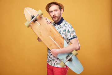 Surpised young man with handlebar mustache dressed in tropical print polo shirt, red shorts and straw hat, with longboard in his hands. Studio shot with yellow background