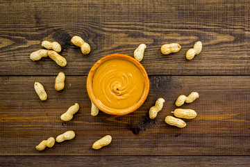 Product for hearty breakfast. High-calorie product. Peanut butter in bowl near nuts in shell on dark wooden background top view