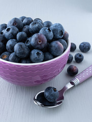 Bright purple bowl full of fresh ripe blueberries, few blueberries on the side, tea spoon with lilac  polka dot handle