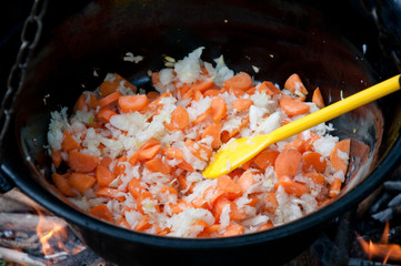 delicious meal is frying of onions and carrots in big metal kettle, with yellow plastic spoon  outdoor close up