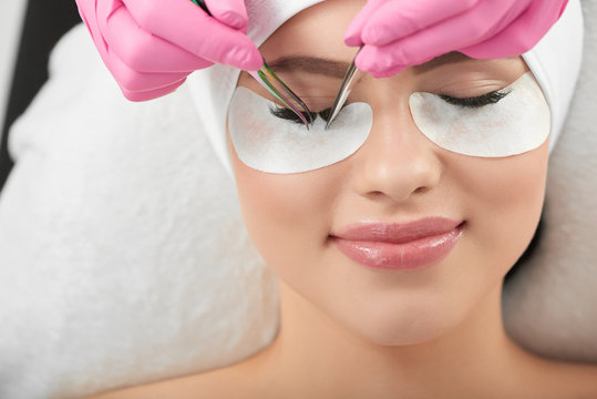Professional photo of smiling model lying during lasmkaing process. Making long lashes for a client wearing pink hygienic gloves. Master is using different types of tweezers. Girl with closed eyes.