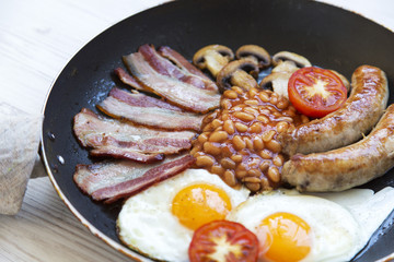 Full English breakfast in cooking pan with fried eggs, bacon, sausages, beans and toasts. Closeup.