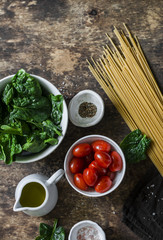 Ingredients for vegetarian healthy lunch - whole grain spaghetti pasta, cherry tomatoes, fresh spinach on a wooden background, top view. Flat lay, copy space
