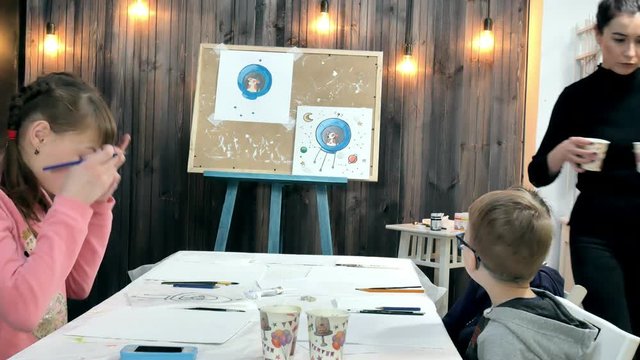 Children boys and girls sitting together around the table in classroom and drawing. With them is their young and beautiful teacher.