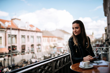 Daydreaming. Lovely smiling girl sitting in the cafe on a balcony over the european city street.