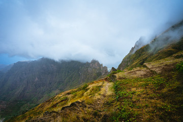 Fototapeta na wymiar Trekking path beside the mountain peak chain overgrown with verdant grass and cultivated plants. Foggy clouds moving over the Mountain edge. Xo-Xo Valley. Santo Antao Island, Cape Verde Cabo Verde
