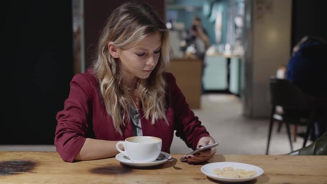 Beautiful young woman with blonde curly hair drinking coffee in a cafe and looking at her smartphone screen. Concept of a business lifestyle. Left to right pan real time medium shot