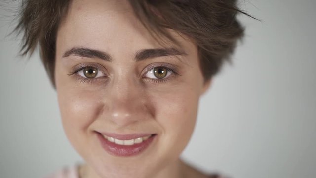 Beautiful teenage girl with short brown hair wearing a pink t shirt thinking and blinking while standing against a gray wall. Handheld slow motion close up shot