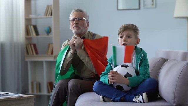 Grandfather and grandson waving Italian flags, watching football together