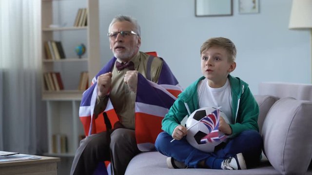 Grandpa and grandson waving British flags, watching football together at home