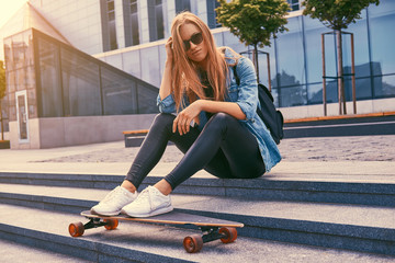Young hipster blonde girl in casual clothes and sunglasses, sitting on steps against a skyscraper, resting after riding on the skateboard.