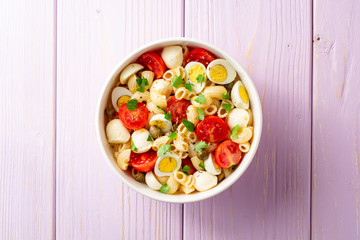 Pasta salad with quail eggs, mozzarella, cherry tomatoes and capers in bowl on purple wooden background