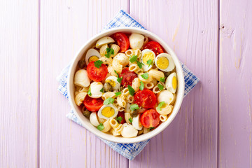 Pasta salad with quail eggs, mozzarella, cherry tomatoes and capers in bowl on purple wooden background