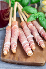 Grissini bread sticks coiled with prosciutto ham on wooden board, vertical
