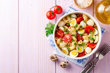 Pasta salad with quail eggs, mozzarella, cherry tomatoes and capers in bowl on purple wooden background