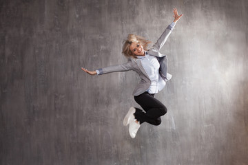 Young blonde woman in business suit and sneakers jumping for joy, gray textured background
