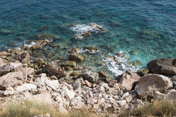 Fototapeta na wymiar Sea rocky coast with clear water of emerald color and white foam on a sunny day, top view; Blue-green sea water and big rose stones on the seashore. 