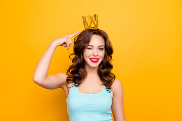 Portrait of cheerful charming girl showing gold crown on head with forefinger winking with one eye looking at camera isolated on yellow background