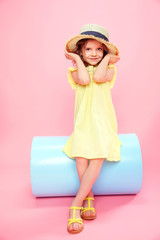 Adorable little girl in yellow light dress and sandals wearing straw hat posing on pink background.
