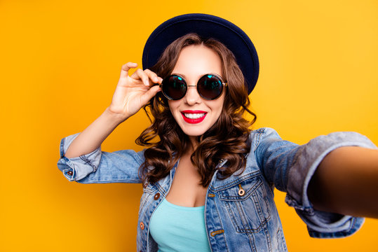Portrait of trendy pretty tourist with beaming smile shooting self portrait on front camera holding eyelet of glasses on face wearing jeans jacket isolated on yellow background, travel tourism concept