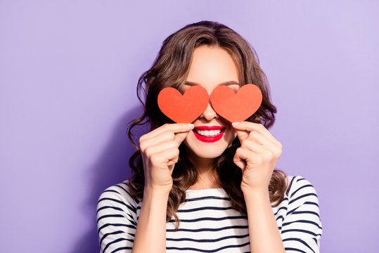 Portrait of lovely creative girl with white teeth red pomade covering closing eyes with two carton paper small little heart figures love signs isolated on violet background