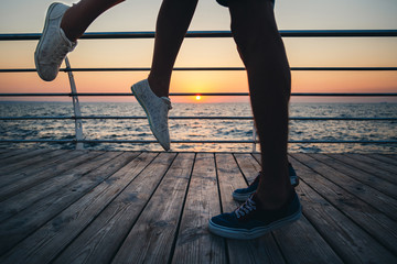 Couple of young hipster lovers kissing, close up on legs and sneakers at the beach at sunrise sky at wooden deck summer time