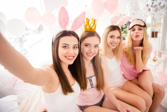 Self portrait of cheerful, pretty, attractive, charming, cute, sexy girls with bunny ears, crown, eye-mask on head shooting selfie on front camera, sending air kiss with palm, enjoying meeting indoor