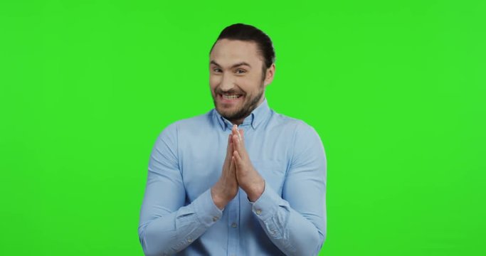 Handsome cheerful young man in the blue shirt standing on the green screen background, smiling archly while rubbing his hands. Chroma key