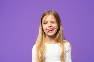 Little girl show tongue. Child with funny face on violet background. Cute kid with beauty look and long blond hair. Beauty look and hair care. Happy childhood and childcare, copy space