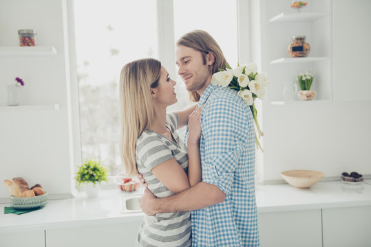 Portrait of sexy modern couple enjoying holiday embracing in kitchen with interior holding bouquet of white tulips in hand kissing looking at each other